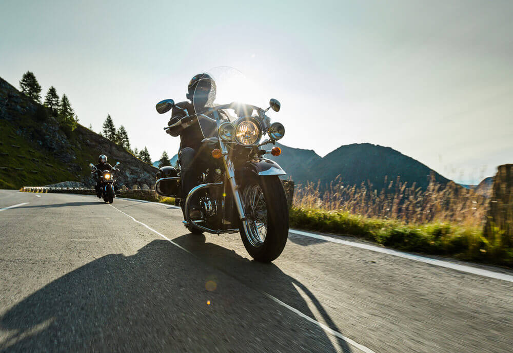 Two motorcyclists riding on a mountain road under a clear sky, with the sun shining brightly.